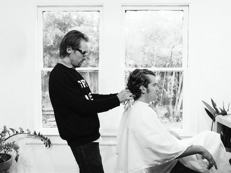 Barber cutting a client’s hair in a bright barbershop near a window.